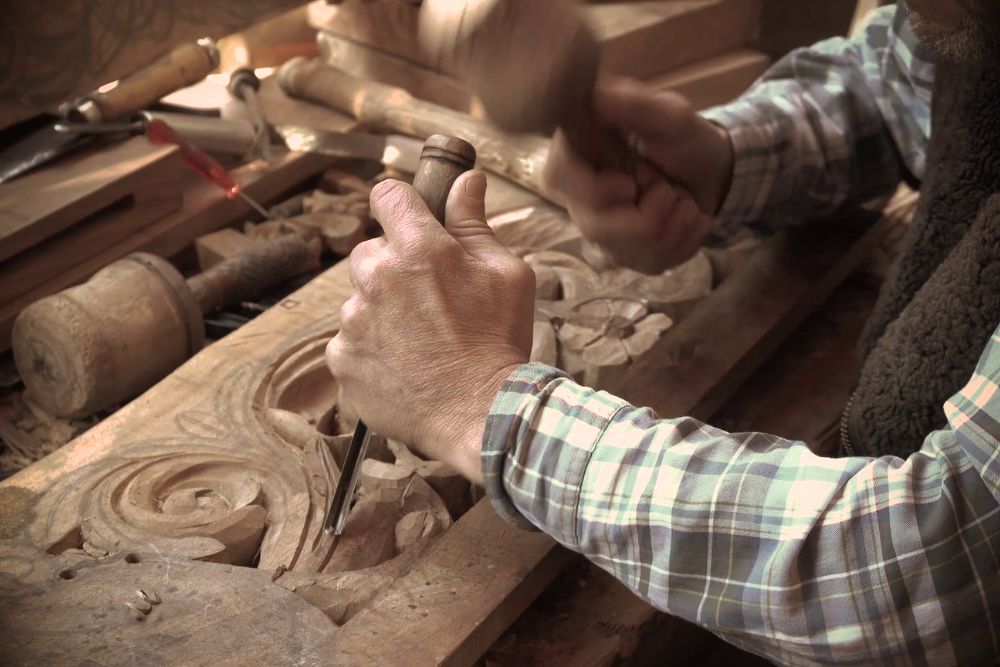 A man focused on intricately carving a piece of wood, surrounded by tools and shavings in a well-lit workshop.