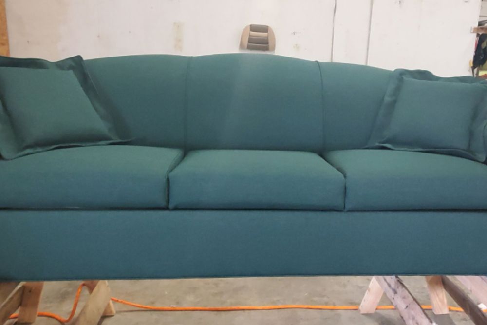 A teal green couch with three cushions and two matching pillows is being crafted in a factory environment.
