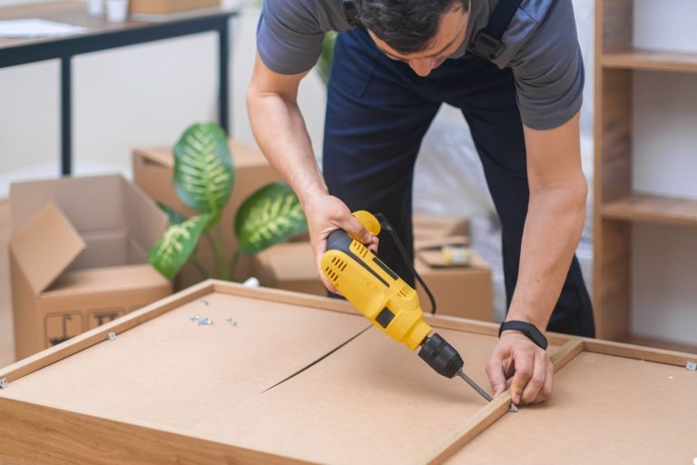 A man operates a drill while constructing a piece of furniture in a workshop setting.