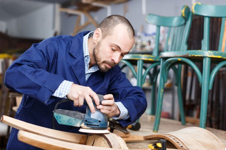 A man using a sander on wood in a workshop, with various tools and wood materials visible in the background.