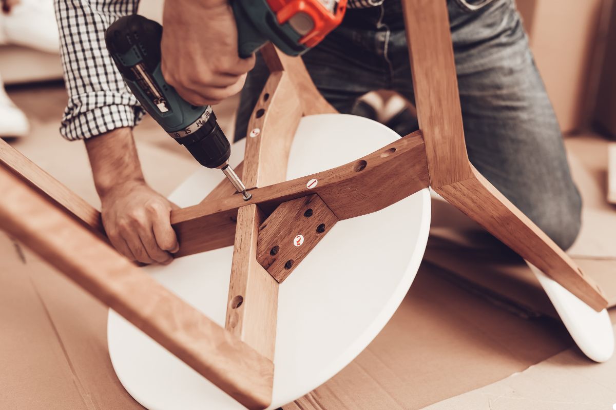 A man operates a drill while assembling a wooden chair in a workshop setting.